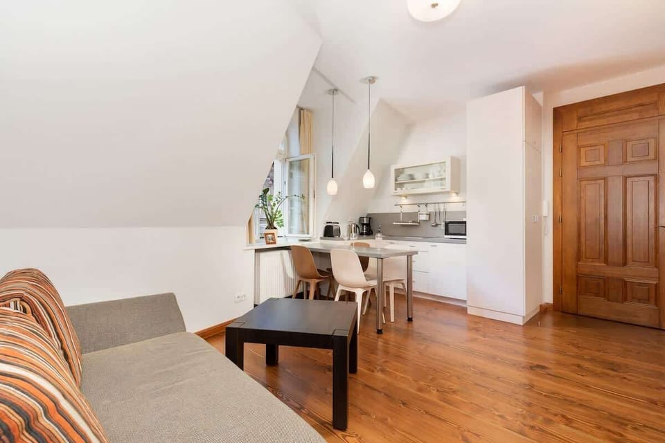 The living room combined with the kitchenette has a cozy, modern vibe. An exposed brick wall adds an industrial touch to the space, while bright furniture and a white dining table lighten up the room. A neutral-colored sofa and a wall-mounted TV make
