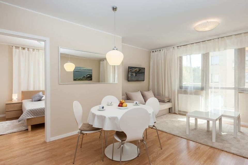 The living room features a soft beige sofa, a white coffee table, and a dining table with modern chairs. Large windows let in natural light, making the room feel open and inviting.