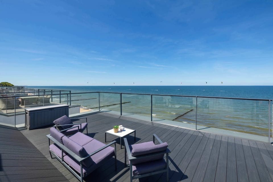 An expansive rooftop terrace is furnished with a comfortable purple outdoor sofa set arranged on dark decking planks. The glass perimeter railing offers an unobstructed view of the tranquil sea under a bright blue sky.