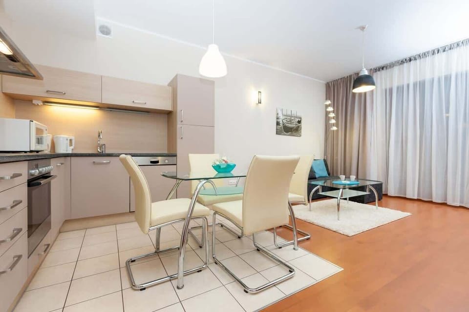 A dining area with a glass table, white chairs, and an open-plan kitchen in the background.
