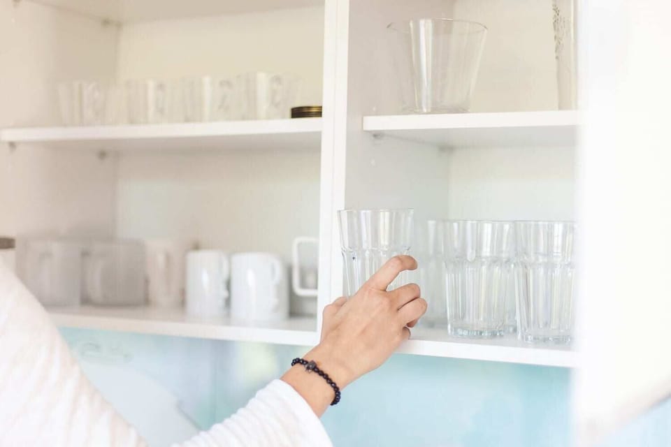  A close-up of a person reaching for glassware inside a white kitchen cabinet. The cabinet is organized, with clear glass cups and white plates neatly arranged.