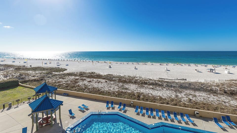 Clear blue pool water sparkles as you watch beachgoers stroll along the white coast sand under a vast sky