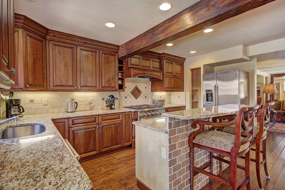 A spacious kitchen with wooden cabinets, granite countertops, stainless steel appliances, and a breakfast bar with high chairs. The kitchen features recessed lighting and a tiled backsplash.