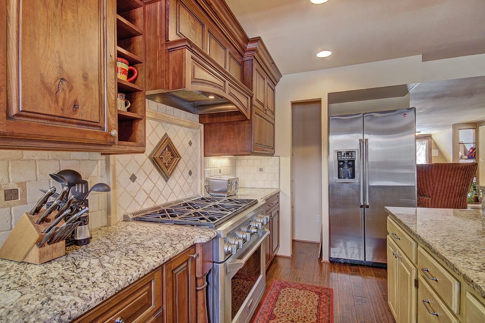A modern kitchen with granite countertops, a stainless steel gas stove, a refrigerator, wooden cabinets, and a small red area rug on the hardwood floor.