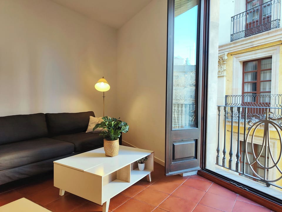 The space is bathed in natural light streaming in through large windows that open onto a charming balcony overlooking the vibrant streets of Barcelona.