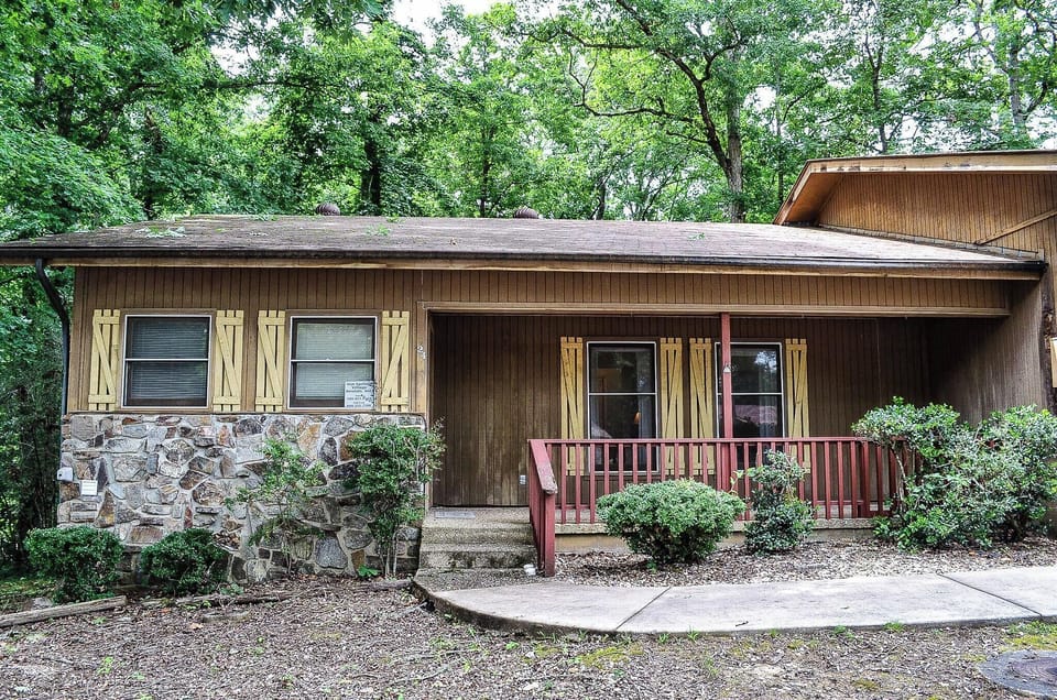 A charming wood-sided cabin with stone accents and a welcoming red porch, nestled in a lush, tree-lined setting that offers peaceful seclusion and natural beauty.