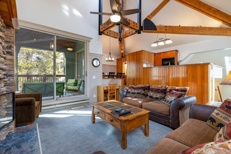 A spacious living room with exposed wooden beams, two leather sofas, a wooden coffee table, a fireplace, and a sliding door leading to an outdoor deck with seating. A kitchen area is visible in the background.