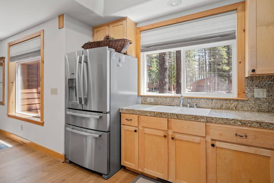 A bright kitchen space featuring granite counters, blonde wood cabinetry, and views of towering pines—perfect for cooking meals together during your forest getaway.