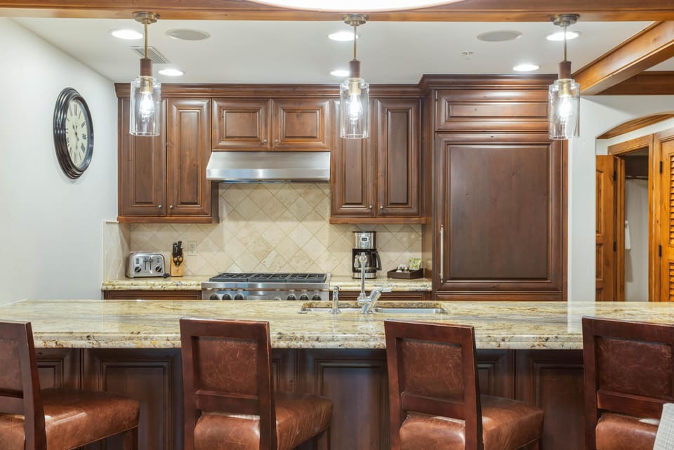 Leather barstools line the granite breakfast bar beneath pendant lights and dark wood kitchen cabinets.