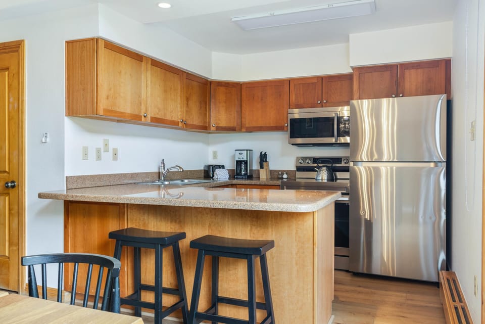Light wood cabinetry and granite countertops surround stainless steel appliances in the kitchen with black barstools at the peninsula.