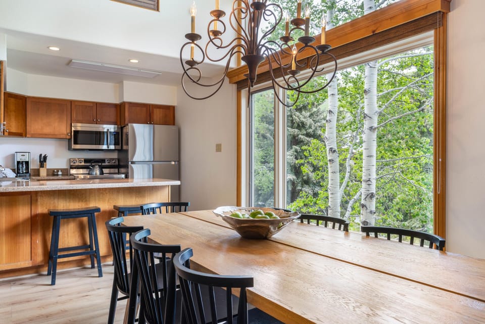 Dining area with a long wooden table, black chairs, and a large window framing aspen trees outside.