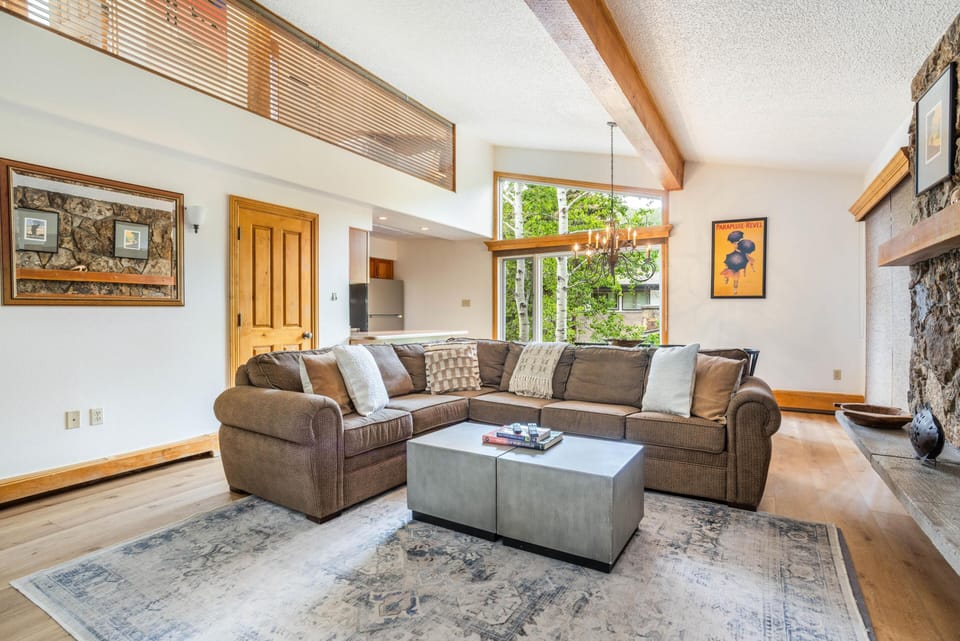 Living room with brown sectional sofa, concrete coffee tables, large windows, and stone fireplace mantel.