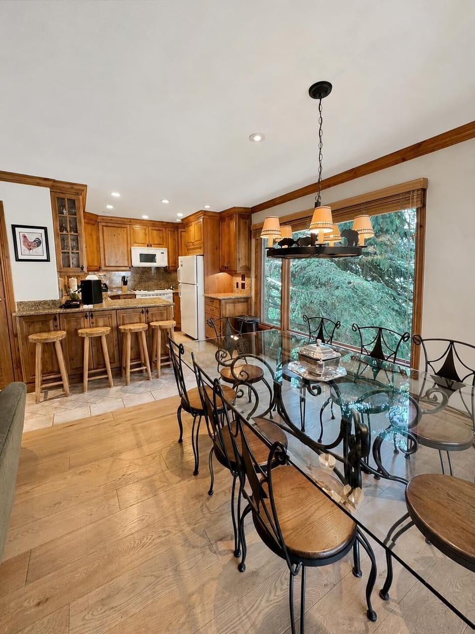 Wrought iron and wood chairs surround a glass dining table beneath a black chandelier by a large window.