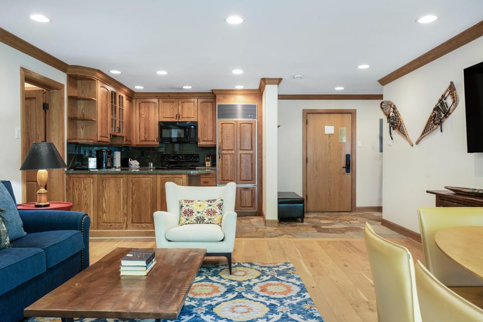 Open living area with hardwood floors, a blue sofa, patterned rug, and wood kitchen cabinets under recessed lighting.