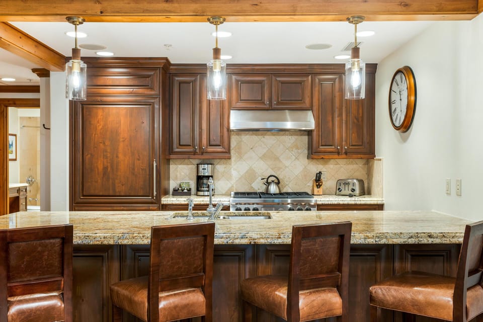 Granite countertop island with built-in sink and four leather-upholstered bar stools under pendant lights.