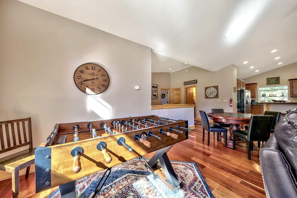 Sunlight highlights the polished hardwood floor beneath a wood-paneled foosball table and round dining table with black leather chairs.