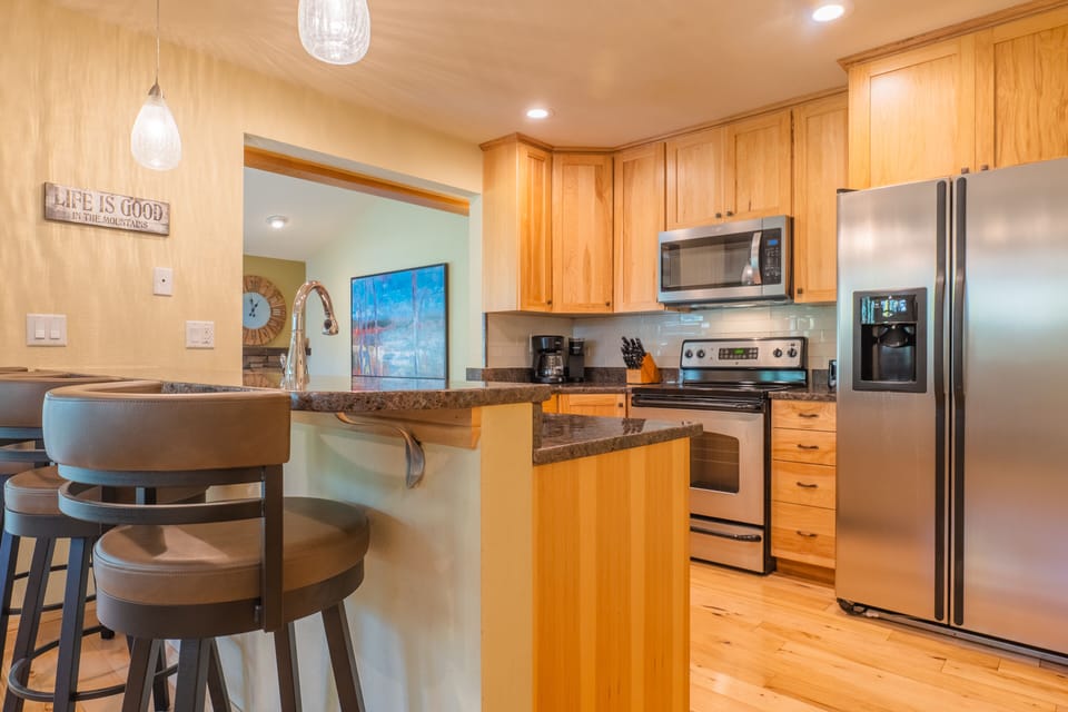 Kitchen with wooden cabinets, stainless steel appliances, and a breakfast bar with three brown padded bar stools. Pendant lights hang above the counter.