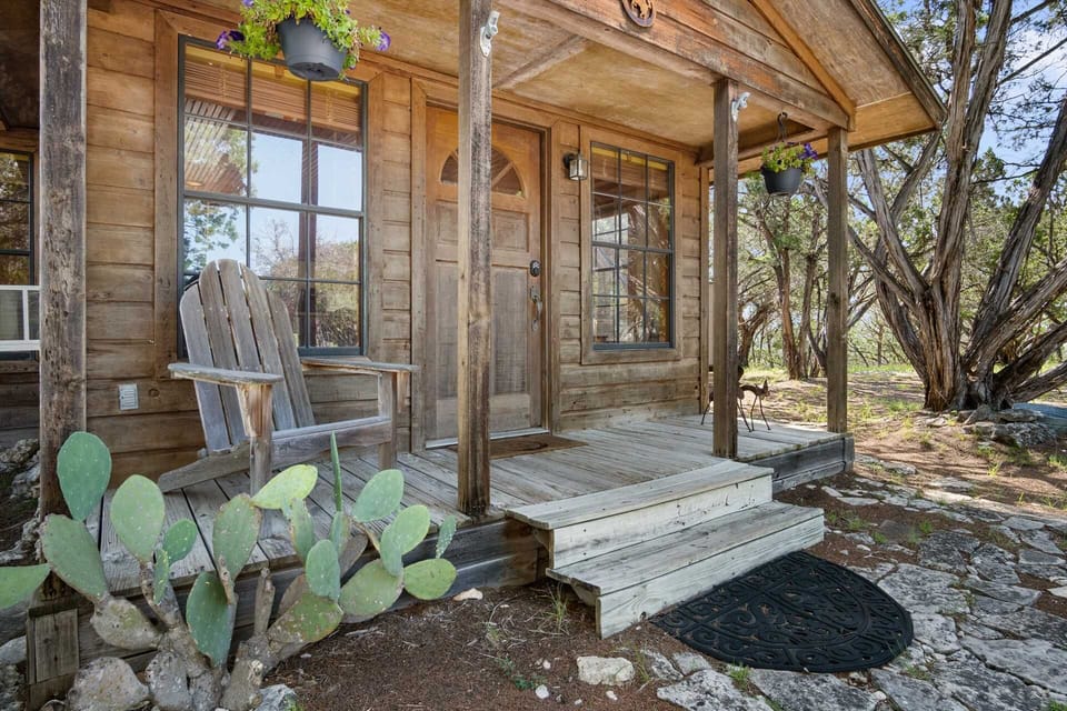 A welcoming wooden cabin porch with an Adirondack chair, blooming hanging baskets, and a cactus plant, surrounded by nature.