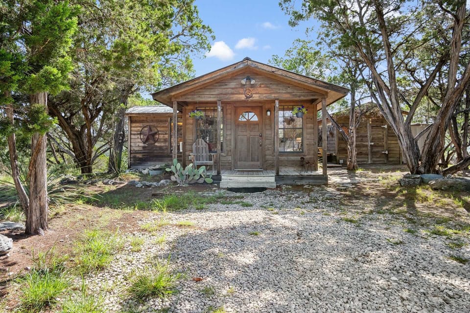 A charming wooden cabin with a small porch is surrounded by trees and a gravel driveway under a bright blue sky.