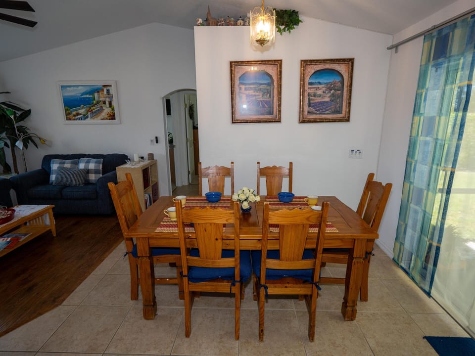 Wood dining table set with six chairs and blue cushions, placed on beige tile next to sheer blue curtains.