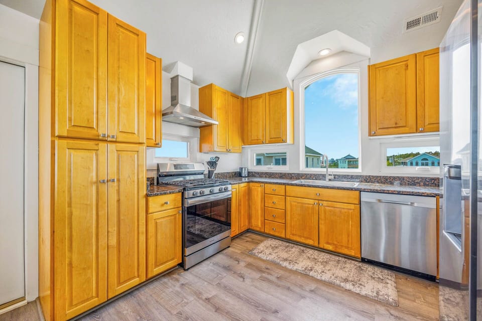 Step into your fully-equipped kitchen with warm wood cabinetry, granite counters, and stainless appliances. Natural light streams through a large window overlooking cheerful neighboring cottages, setting the perfect scene for preparing your favorite meals.