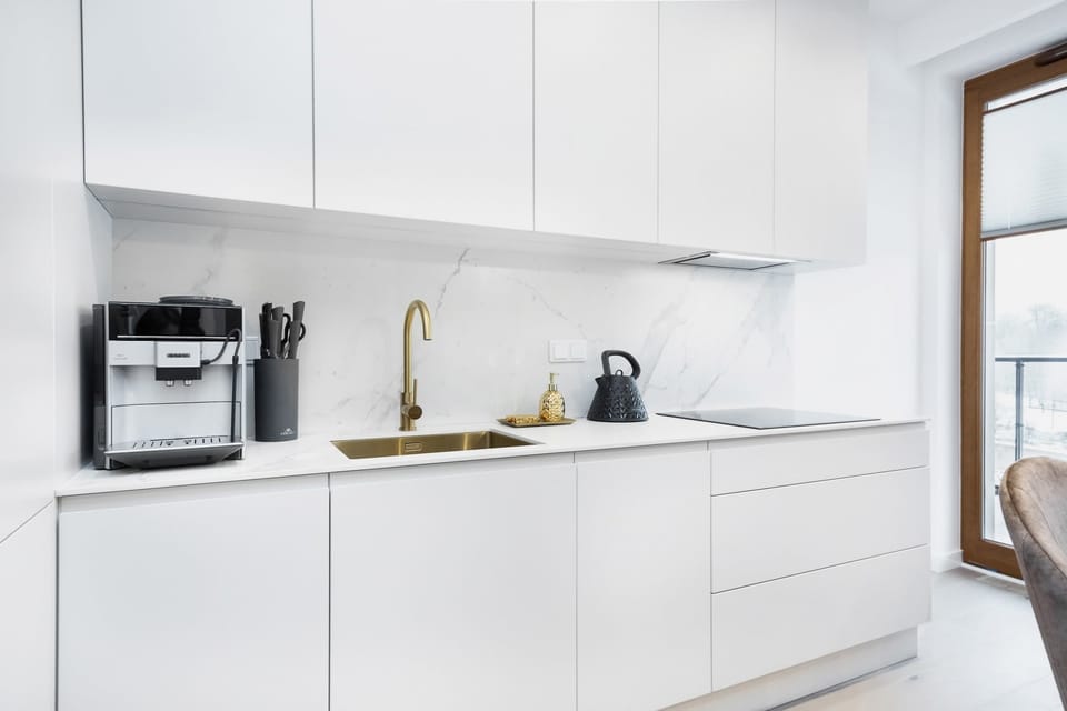 A minimalist kitchen with all-white cabinets and countertops. The setup includes a sink, stovetop, and a coffee machine, emphasizing functionality and cleanliness.