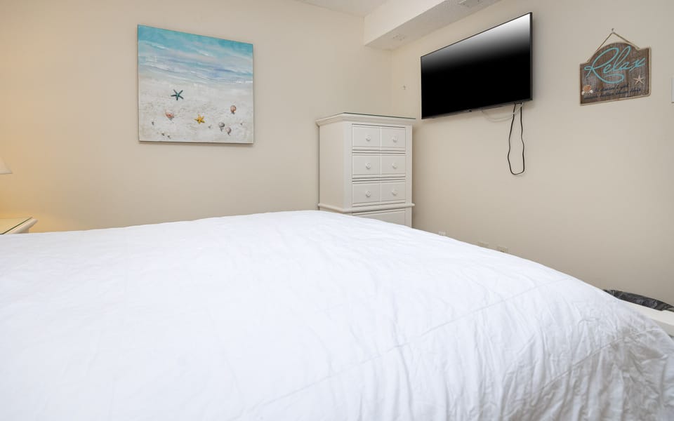 This bedroom features a white quilted bedspread, a mounted TV, and beach-themed artwork for a relaxing atmosphere
