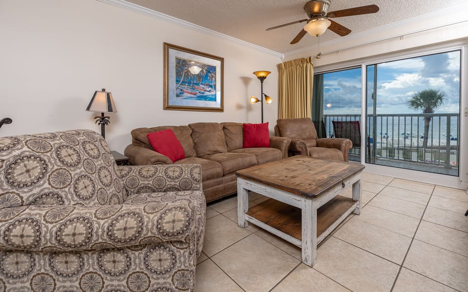 The living area features comfortable seating, a rustic coffee table, and a stunning beach view through the large balcony door