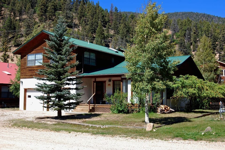 A charming mountain retreat with green roof and wood-sided upper level sits surrounded by evergreens and aspens, with forested slopes rising behind—a peaceful arrival setting.