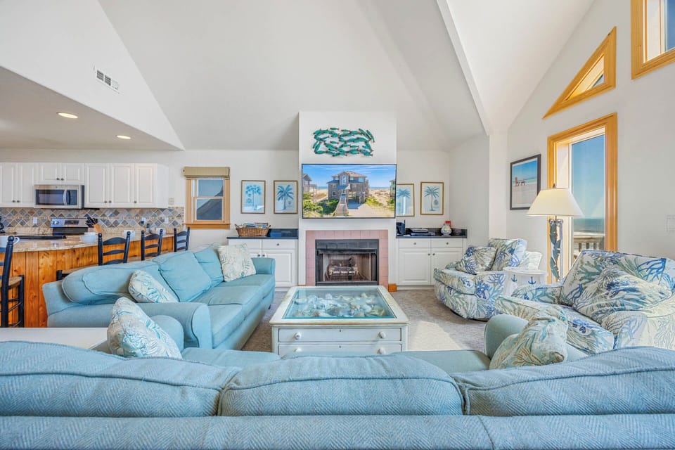 Your spacious coastal living room awaits with vaulted ceilings, plush seating, and a cozy fireplace. Natural light pours through framed windows revealing stunning ocean views that make every moment feel like a beach escape.