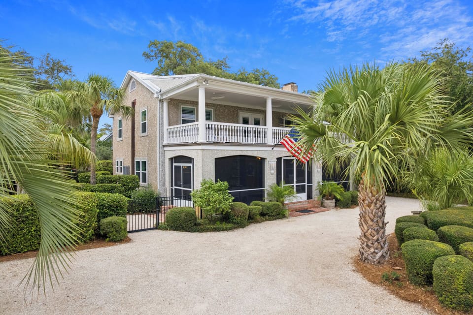 This Southern-style home features a welcoming covered porch, lush landscaping with palms and manicured hedges, and a convenient driveway—offering a charming coastal retreat in a beautifully maintained setting.