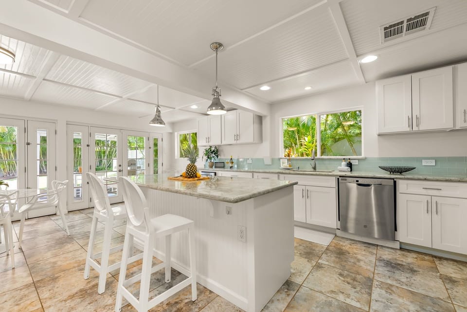 Step into your bright, coastal-inspired kitchen with white cabinetry, granite counters, and stainless appliances. Large windows frame lush palm views, flooding the space with natural light.