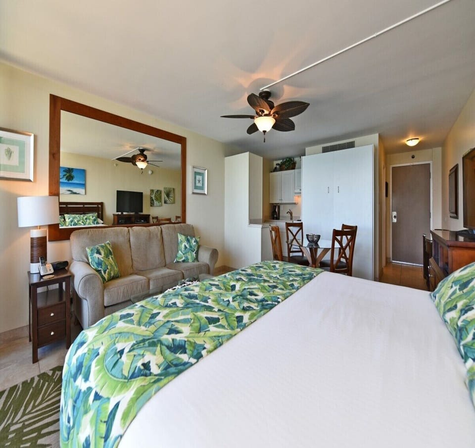 Room angle highlighting beige sofa with matching leaf‑patterned cushions, large mirror above, bed and ceiling fan visible, dining area and kitchenette completing the tropical setting.