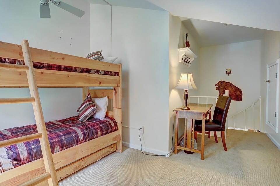 A room with a wooden bunk bed, a small desk with a lamp, a chair, and a decorative wooden bear on the wall. Stairs and a railing are visible in the background.