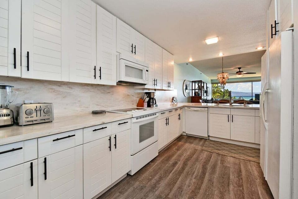 Modern kitchen with white cabinetry, stainless appliances, and wood‑style flooring, chandelier lighting the dining area beyond.