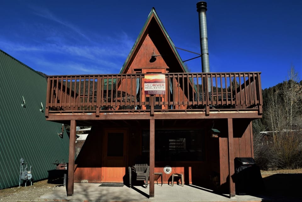 Rustic A-frame mountain retreat surrounded by aspen trees and natural landscape. The property's distinctive architecture stands beneath vivid blue skies in a serene mountain setting.