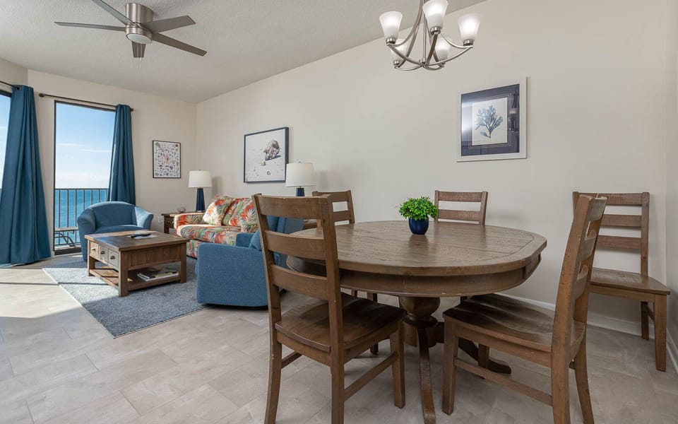 Bright living area features a floral sofa, blue armchairs, and a dining table, with expansive views of the beach beyond
