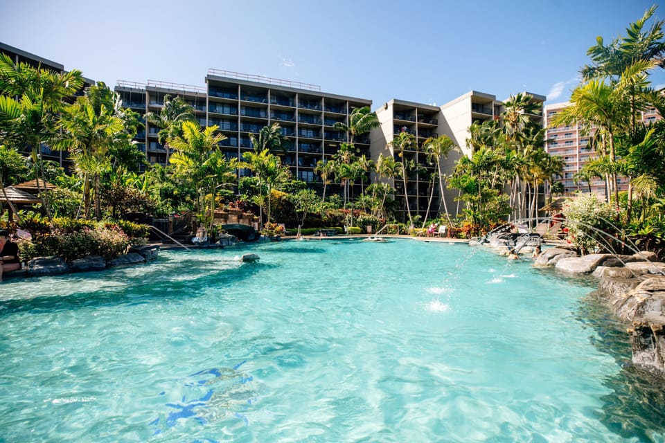 The property features a stunning tropical pool area with crystal-clear waters, lush landscaping, and palm trees creating a resort-style oasis beneath sunny skies.