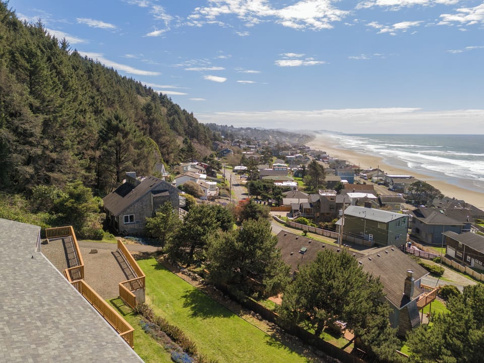 Elevated coastal community nestled between forested hills and pristine sandy beach with rolling ocean waves stretching to the horizon.