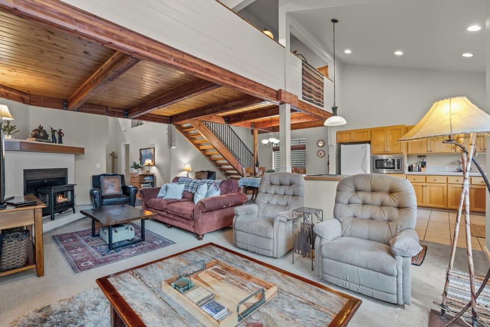 Soaring ceilings with rich wooden beams frame this open-concept living space where you can unwind by the cozy fireplace, gather with loved ones, and enjoy seamless flow to your fully equipped kitchen.
