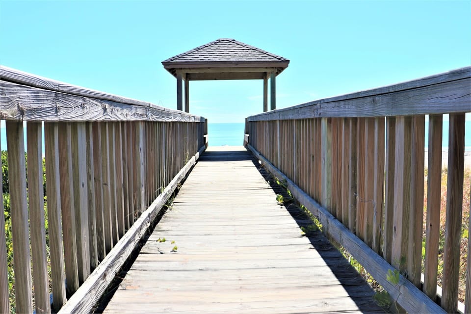A wooden boardwalk extends toward a covered pavilion and the ocean beyond, offering direct beach access from the property.