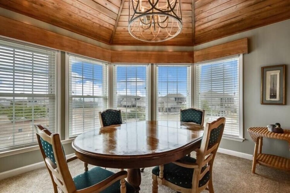 Gather around the elegant wooden dining table under a rustic chandelier, where natural light pours through expansive windows and vaulted wood ceilings create a warm, inviting atmosphere for memorable meals together.