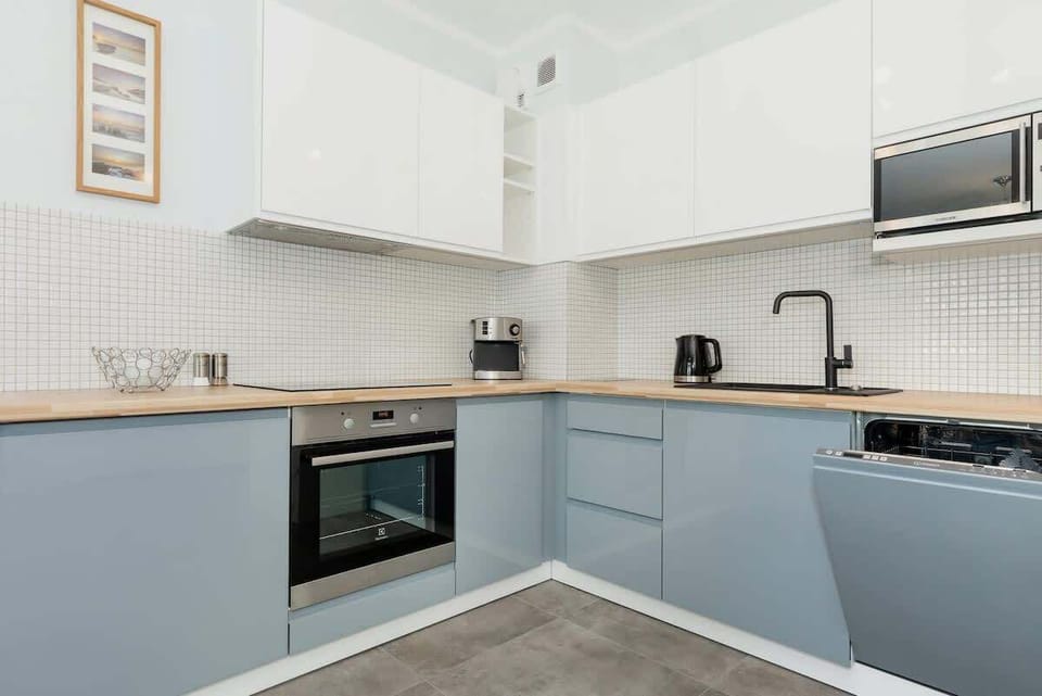 A compact modern kitchen with white cabinets, a tiled backsplash, and stainless steel appliances. The flooring is light gray.