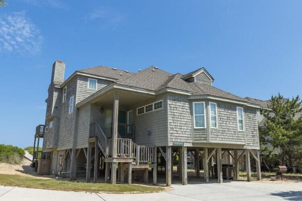 Coastal vacation home elevated on stilts with classic shingle siding, featuring covered parking and multiple outdoor areas on the surrounding dunes.