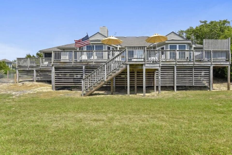 A charming beachfront retreat elevated on stilts with spacious wraparound decks, surrounded by open lawn and coastal greenery under clear blue skies.