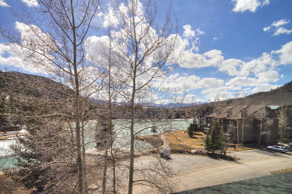 A scenic landscape featuring bare trees, a parking lot, a few buildings, and a river with mountains in the background under a partly cloudy sky.