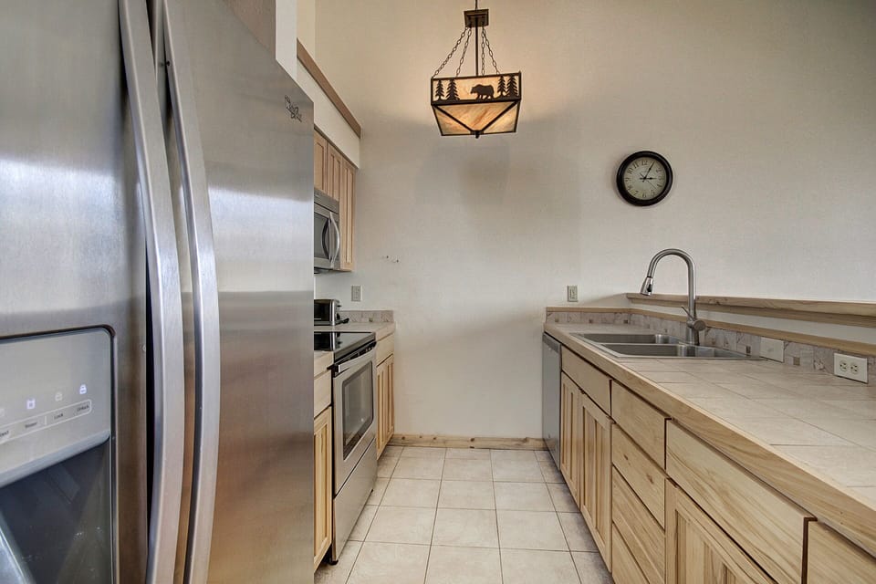 A modern kitchen with stainless steel refrigerator, oven, microwave, light-colored wooden cabinets, tiled countertops and floors, a clock on the wall, and a ceiling light featuring a bear design.