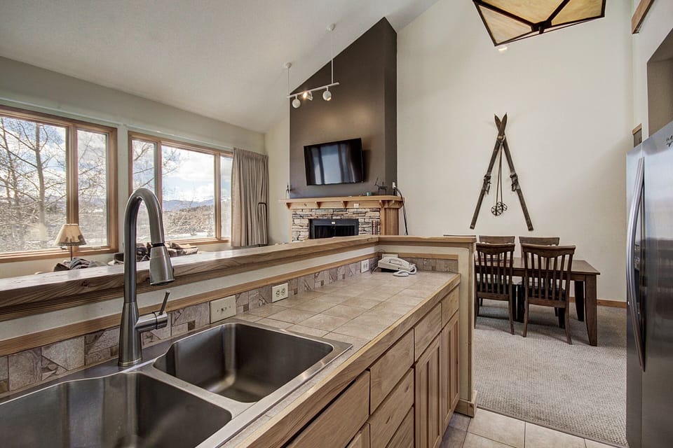 Kitchen area overlooking a dining and living room with a high ceiling, skylight, and mounted skis on the wall. A large window and a wall-mounted TV above the fireplace are visible.