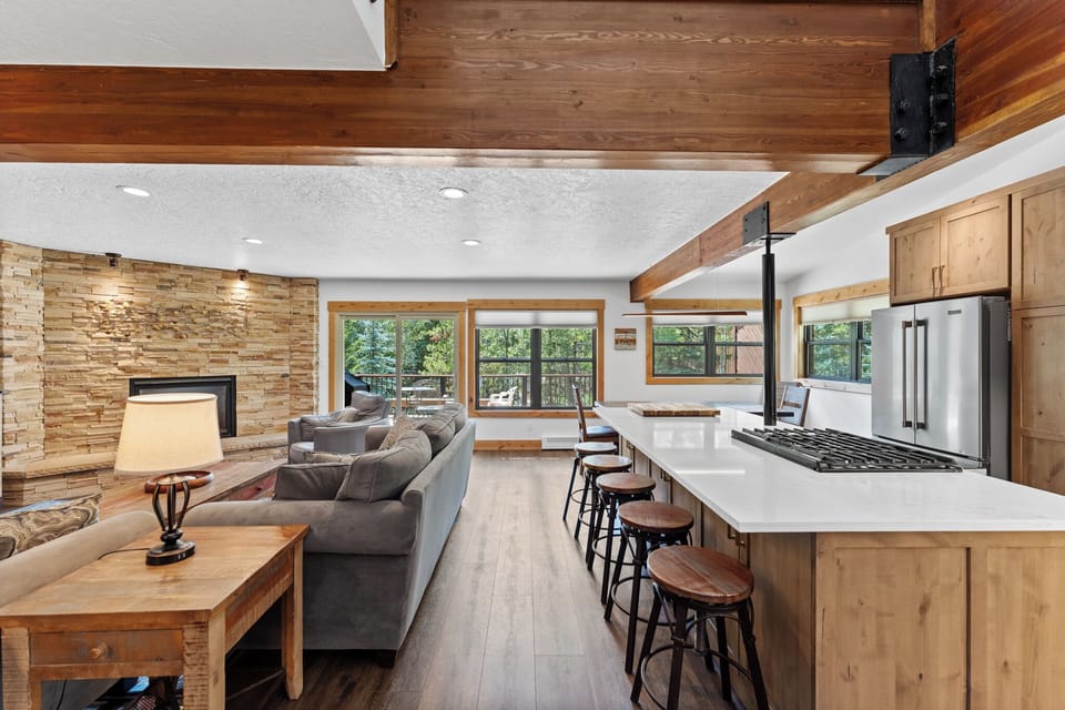 Relax in your spacious open-concept living area featuring exposed beams, a stone fireplace, and modern kitchen with breakfast bar seating.