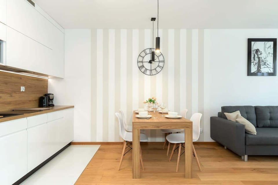 A compact kitchen and dining area with white cabinetry and a small dining table. A black accent clock and pendant lights add style.